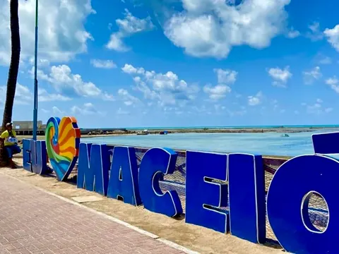 Letreiro azul Eu amo Maceió na orla, com o mar e o céu azul ao fundo