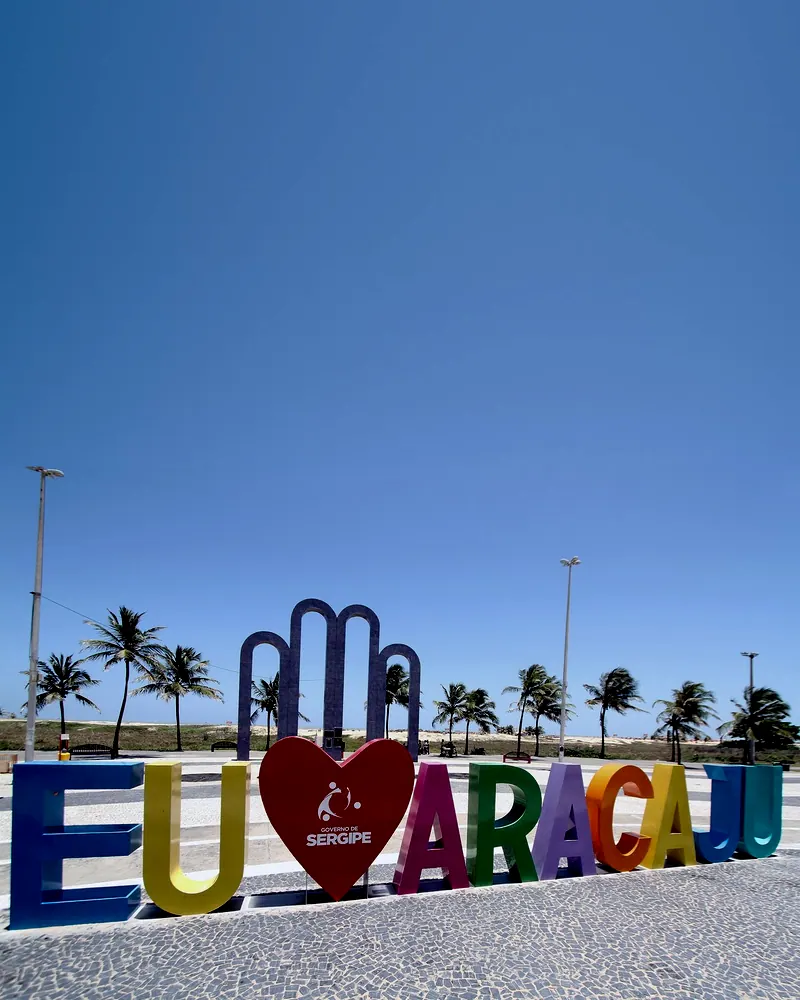 Letreiro colorido Eu amo Aracaju inteiro na Orla de Atalaia, com os Arcos da Orla e coqueiros ao fundo